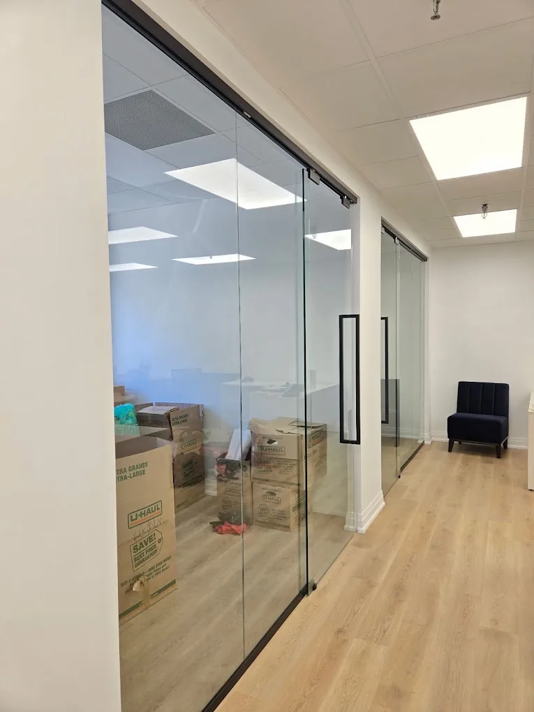 Interior corridor with glass partitions, black-framed doors, ceiling lighting, and wood flooring